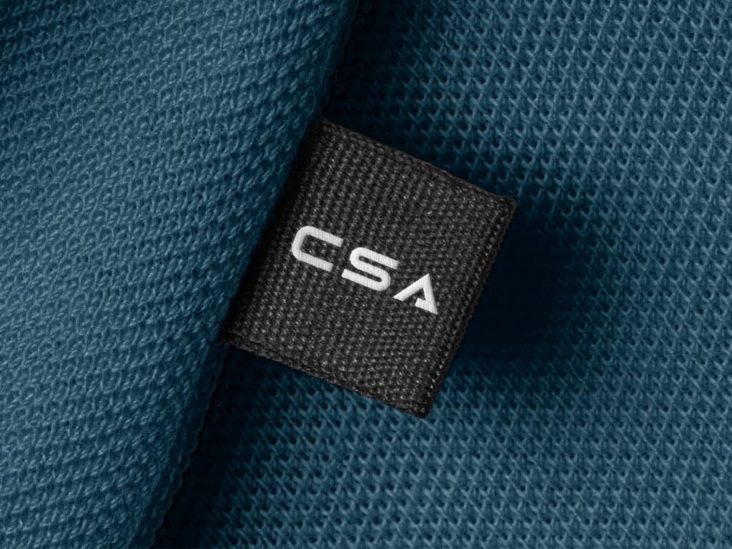 Zwart geweven label met witte letters ‘CSA’, bevestigd op een textielproduct. Minimalistische branding met industriële uitstraling.