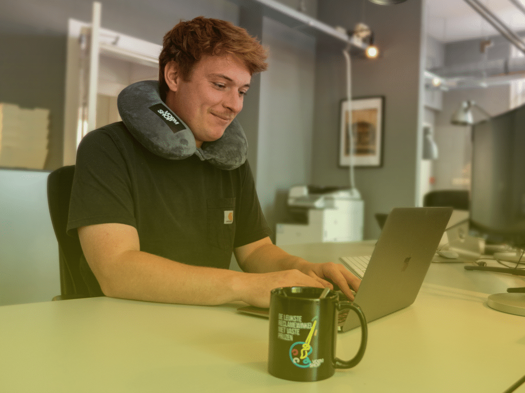 Man werkt op laptop aan bureau met nekkussen tijdens werkmoment op kantoor.