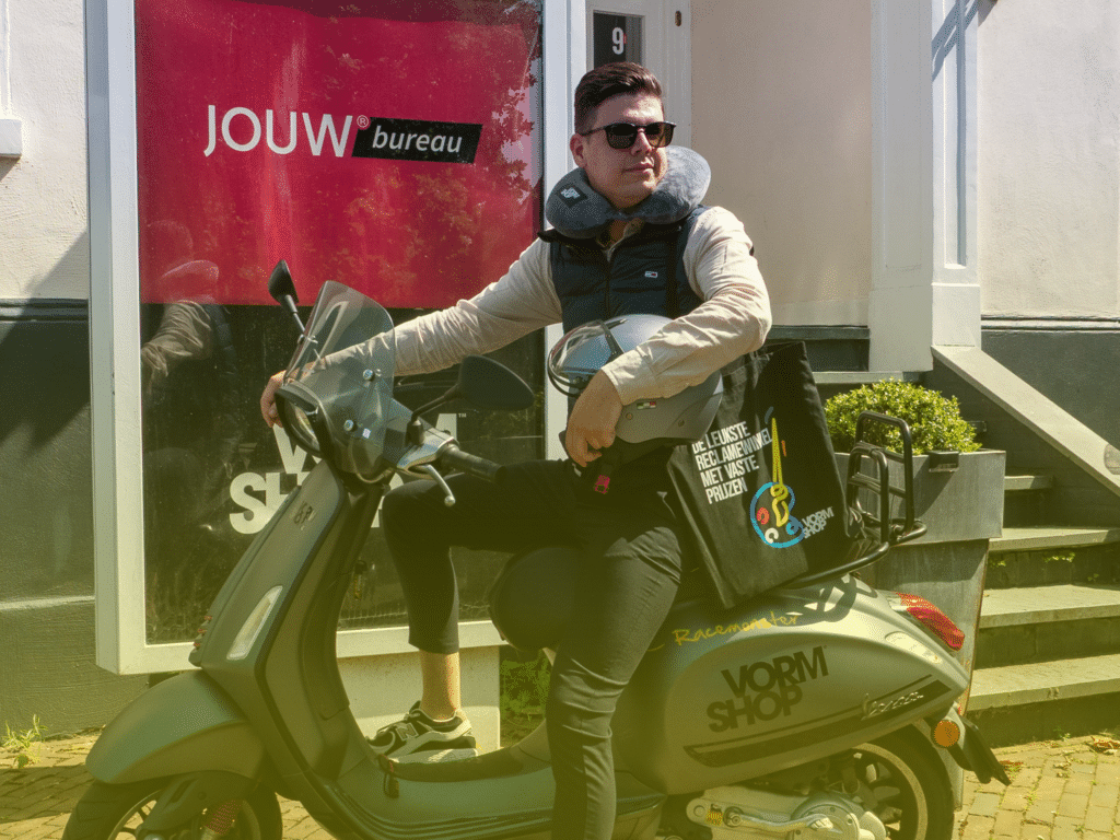 Man zit op scooter voor kantoor van JOUW bureau / VORMSHOP met nekkussen om zijn nek.